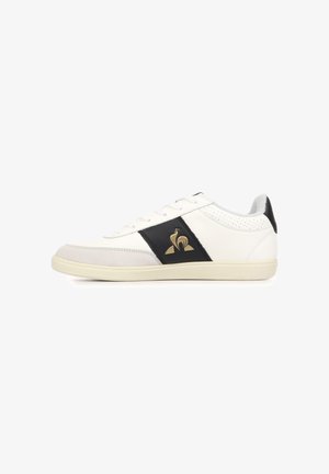 Sneaker bianca bassa con pannello laterale nero con logo gallo dorato, lacci bianchi e suola beige chiaro.