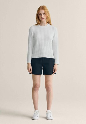Kvinde med rødt hår iført tekstureret hvid langærmet sweater, sorte shorts og hvide sneakers, stående mod en ensfarvet beige baggrund.