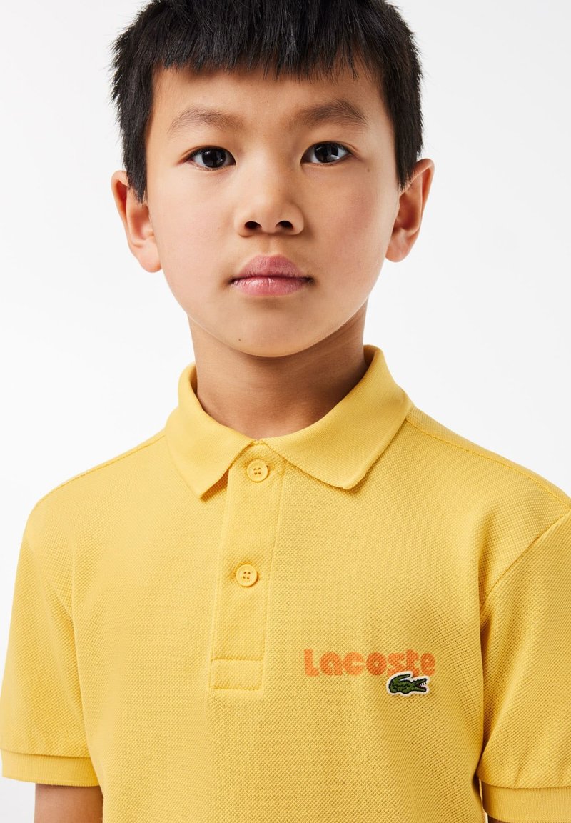 Polo jaune en tissu texturé, avec un col à trois boutons et un logo "Lacoste" brodé avec un accent crocodile vert.