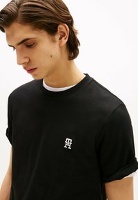 Czarny bawełniany t-shirt z okrągłym dekoltem i krótkimi, podwiniętymi rękawami. Z przodu znajduje się małe białe logo na lewej piersi. Gładka tekstura, casualowy styl.