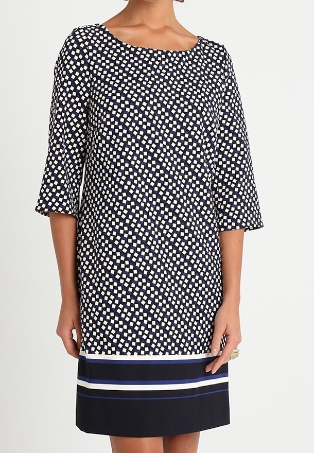 Femme portant une robe mi-longue à manches trois-quarts, avec un motif géométrique noir et blanc et un ourlet rayé bleu marine.