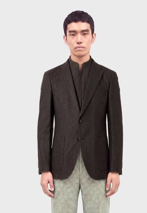 Homme portant un blazer marron foncé sur mesure par-dessus un haut à fermeture éclair avec un pantalon à carreaux clair, debout devant un fond uni.