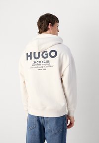 Bijela dukserica s velikim crnim "HUGO" logom na leđima, s mekim materijalom i kapuljačom s vezicama. Uparena s plavim trapericama.