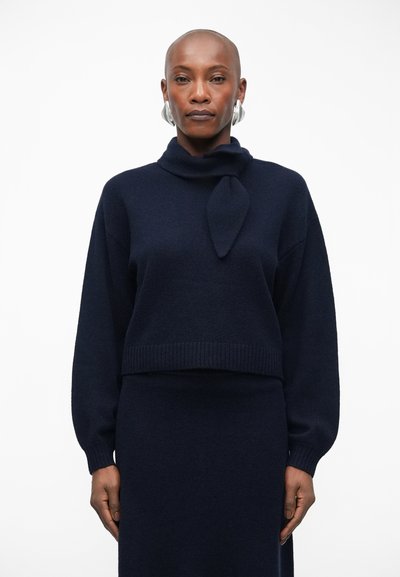 Pull en maille bleu marine avec col replié et détail de nœud à l'avant, associé à une jupe assortie. Texture douce et accents à ourlet côtelé.