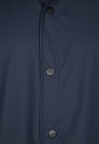 Navy vesikäypär jacket, jossa on suuret mustat painonapit, sileä pinta ja hienovaraiset ommel-yksityiskohdat placketin varrella.