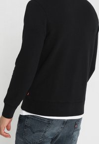 Personne portant un sweat-shirt noir par-dessus une chemise blanche avec un jean en denim bleu, vue de dos, de la taille jusqu'aux épaules.