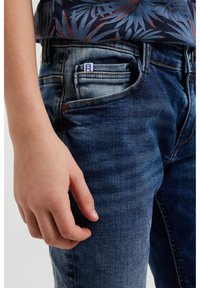 Dž ínové kalhoty v tmavě modré barvě s mírným efektem vyblednutí. Mezi rysy patří přední kapsa a viditelná značka. Ruka odložená na noze.