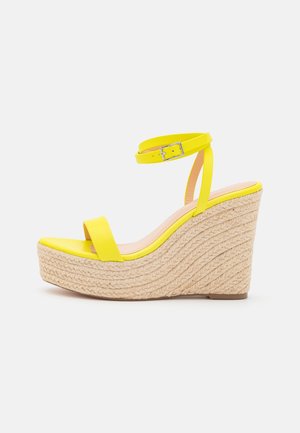 Sandale compensée jaune avec plateforme en corde tressée, bride ouverte sur les orteils et bride de cheville ajustable avec boucle argentée sur fond blanc.