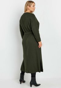 Cardigan long côtelé vert olive avec une taille ceinturée, fentes sur les côtés et manches longues. Fabriqué en matériau texturé ; porté avec des bottines noires à cheville.