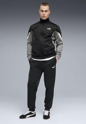 Jonge man staat met handen in zijn zakken, draagt een zwarte en grijze Puma trainingspak en zwarte Puma sneakers, tegen een grijze achtergrond.