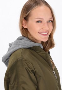 Olíva zöld bomber dzseki szürke kapucnival, cipzárral záródik és bordás mandzsettákkal rendelkezik. Sima tapintású és testhez álló fazonú.