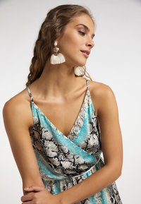 Sukienka bez rękawów, z zakładanym krojem, w niebiesko-czarny wzór wężowy. Modelka nosi kremowe kolczyki z frędzlami. Gładki materiał, dopasowana góra.