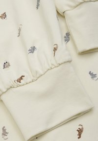 Crème kleurige stof met elastische manchetten, voorzien van kleine dinosauruspaternen in bruin, grijs en blauw over het oppervlak.