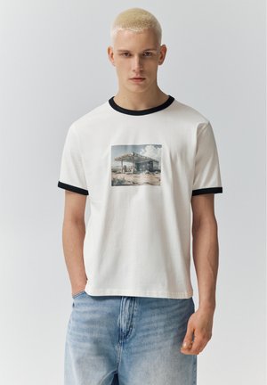 Ung mand med kort blondt hår iført en hvid T-shirt med sort kant og et tankstationsmotiv, kombineret med lyseblå jeans.