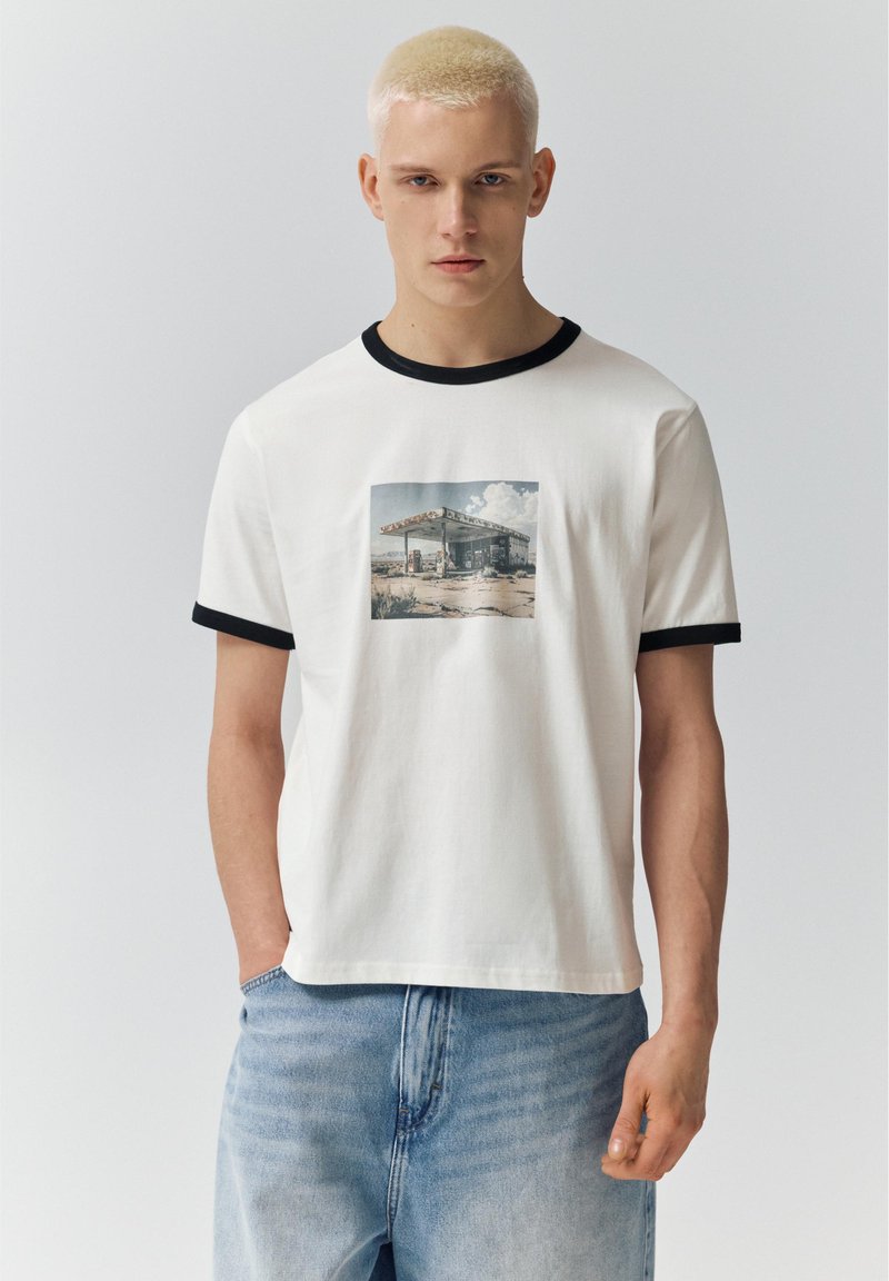 Jeune homme aux cheveux blonds courts portant un t-shirt blanc à bordures noires avec un motif de station-service, associé à un jean bleu clair.