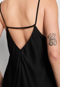 Robe noire à fines bretelles, avec un dos en V profond et des plis subtils. Présente un tatouage géométrique d'ours sur le bras. Tissu lisse et mat.