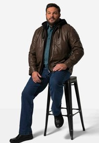 Chaqueta de cuero marrón con capucha negra, llevada sobre una camisa azul de botones, combinada con jeans azules y zapatos negros, sentado en un taburete negro.