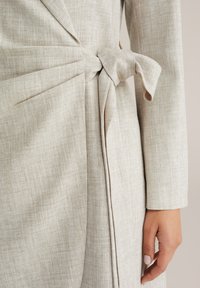 Vestito avvolgente beige chiaro con tessuto strutturato, maniche lunghe e dettaglio con fiocco laterale, che crea una silhouette definita ma rilassata.