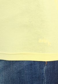 Yellow cotton T-shirt with a round neckline, featuring a stitched hem and subtle logo detail below. Paired with blue denim jeans.