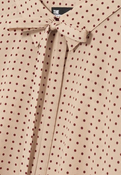 Blusa beige con pois bordeaux, caratterizzata da un fiocco annodato al colletto e un dettaglio del colletto a punta.