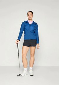 Blå zip-jacka med texturerad framdel, i kombination med svarta shorts och en golfklubba. Modellen har en ljusrosa tröja under och vita sneakers.