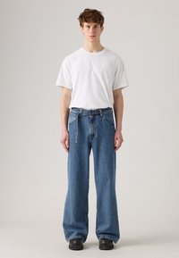 White t-shirt on a person, paired with loose-fitting mid-blue denim jeans featuring a unique belt design and black platform shoes.
