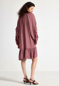 Lång, löst sittande klänning i mauve med volangärmar och samlad nederkant. Slät tyg, knälång, kompletterad med öppna klackar.