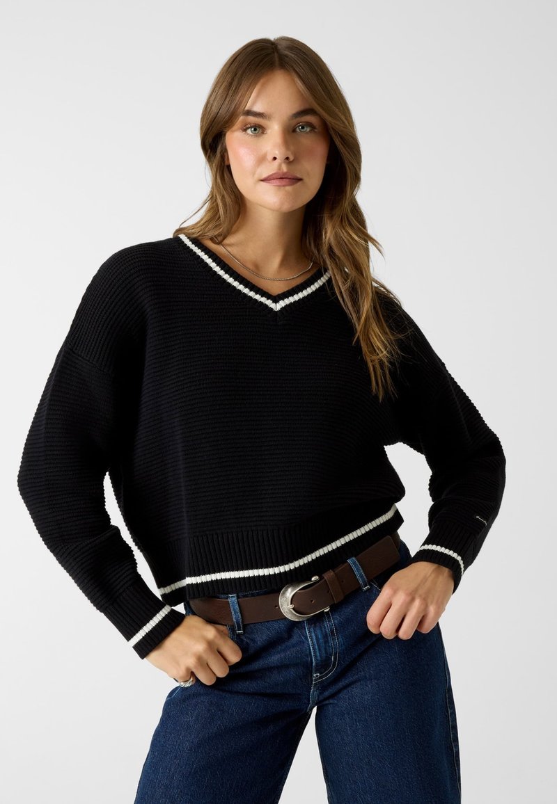 Jeune femme aux longs cheveux bruns portant un pull noir à col en V avec un bord blanc, un jean bleu et une ceinture marron, debout, les mains sur les hanches.