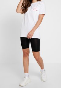 Bílý bavlněný tričko s červeným textem, kombinované s černými cyklistickými šortkami do kolen. Obuv tvoří bílé tenisky s tlustou podrážkou.