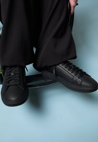 Black leather sneakers with a smooth texture, featuring flat laces and a subtle logo on the side. Set against a light blue background.