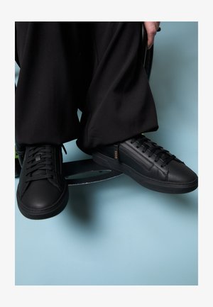 Černé kožené tenisky s hladkým povrchem, s plochými tkaničkami a nenápadným logem na boku. Umístěné na světle modrém pozadí.