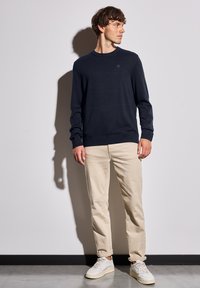 Marinefarbener Strickpullover mit Rundhalsausschnitt, kombiniert mit hellbeigen Chinos und weißen Sneakers, die einen lässigen, taillierten Stil zeigen.