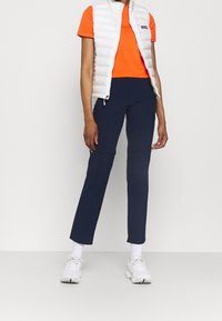 Witte puffy vest over een oranje T-shirt met korte mouwen, gecombineerd met marineblauwe sportbroeken en witte sneakers, met een slanke pasvorm en een gladde textuur.