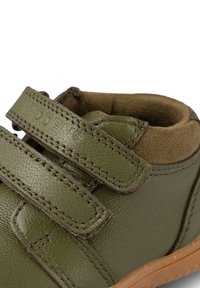 Scarpa verde in pelle per bambini con una finitura testurizzata, dotata di due cinturini in Velcro e di un colletto imbottito marrone. La suola in gomma è di un marrone chiaro.