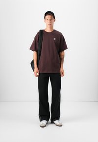 Tmavofialové oversized tričko s bielym logom, doplnené o čierne široké nohavice a biele tenisky. Modelka nosí čiernu tašku.