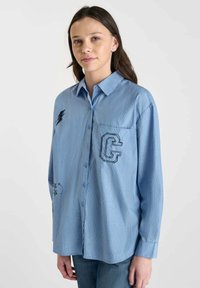 Blue striped button-up shirt with a chest pocket featuring a black "G" logo and embroidered lightning bolt and star designs. Cotton fabric.