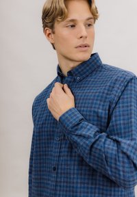 Blå rutete skjorte med knapper og krage, med teksturert stoff og kontrasterende blå knapper, vist i en liten vinkel.