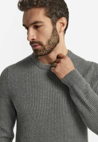 Grå rutad stickad tröja med rund halsringning och texturerade ärmar, visad på en modell som håller i halsringningen.