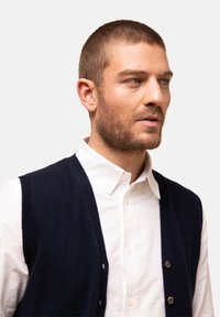 Marinblå stickad väst med V-ringning, texturerat tyg och sex knappar. Bärs över en vit skjorta med krage. Närbildsprofil.