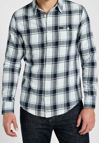 Man som bär en vit och marinblå rutig skjorta med lång ärm och mörka jeans, stående med avslappnade händer.