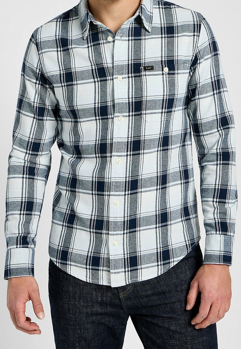 Man som bär en vit och marinblå rutig skjorta med lång ärm och mörka jeans, stående med avslappnade händer.