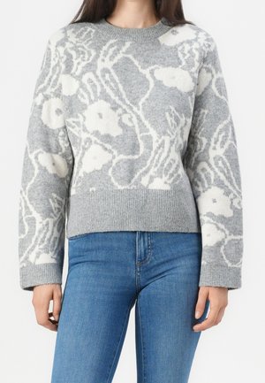 Frau trägt einen grauen Pullover mit weißen Blumenmustern und blaue Jeans, steht vor einem weißen Hintergrund.