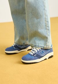 Siniset lenkkarit, joissa on teksturoitu kangaspinta, valkoiset yksityiskohdat ja tukeva musta ulkopohja, yhdistettynä vaaleisiin farkkuihin. Keltainen tausta.