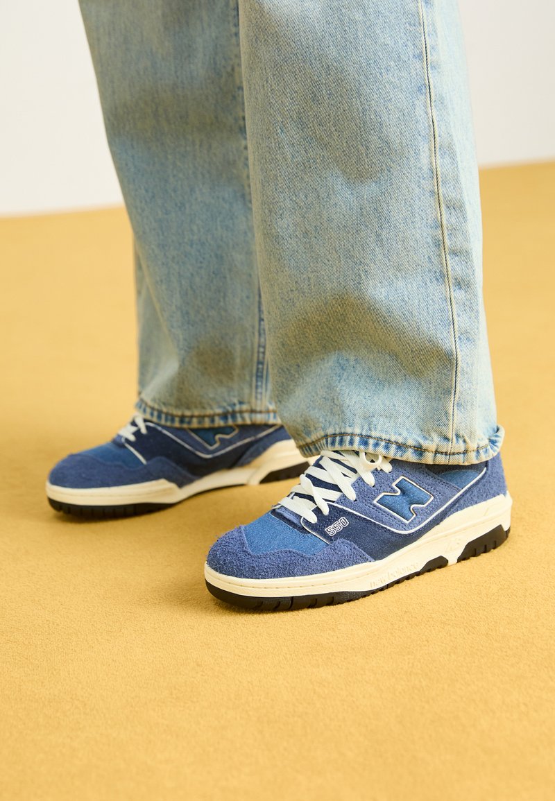 Baskets bleues avec un dessus en tissu texturé, accents blancs et une semelle noire robuste, associées à un jean en denim clair. Fond jaune.
