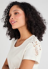 Camiseta de color crema claro con detalles de encaje de ganchillo en los hombros. Tejido suave y escote redondeado. El corte es relajado y casual.