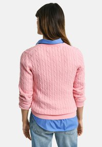 Růžový svetr s pleteným vzorem a dlouhými rukávy, s žebrovanými manžetami, vrstvený přes modrou košili s límečkem, viděný zezadu.