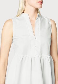 Vestido branco sem mangas com um colarinho alto, apresentando uma frente com pequenos botões e uma cintura franzida para uma silhueta em A.