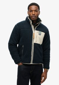 Giacca navy morbida con pannello crema, colletto alto, cerniera integrale e tasche laterali. Presenta un logo del marchio e tessuto texturizzato.