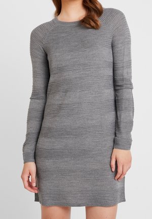 Kobieta ubrana w dopasowaną, długorękawową, szarą dzianinową sukienkę z teksturowanym poziomym prążkowaniem i okrągłym dekoltem, na jednolitym tle.