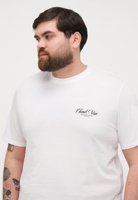 White cotton t-shirt featuring a rounded neckline and short sleeves. Notable black text logo on the left chest area. Smooth texture.